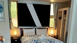 Une chambre moderne avec un grand lit, deux tables de chevet avec des lampes, un fauteuil en osier, une armoire blanche et un placard avec une tringle pour suspendre les vêtements.
