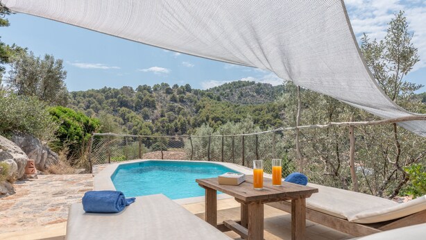 A poolside area with a wooden table, lounge chairs, and a view of a forested hillside.