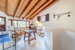 Une pièce avec un haut plafond en bois, un lit recouvert d’une courtepointe à rayures bleues et blanches, deux fauteuils à bascule, une table en bois et une fenêtre donnant sur un paysage verdoyant.