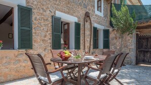 A stone building with a round table and four chairs, a window with a shutter, and a tree.