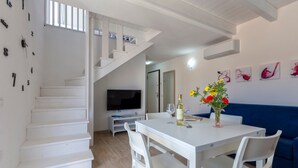 Una escalera blanca que conduce a una plataforma, un comedor con una mesa y sillas, un sofá, y un televisor sobre un mueble.