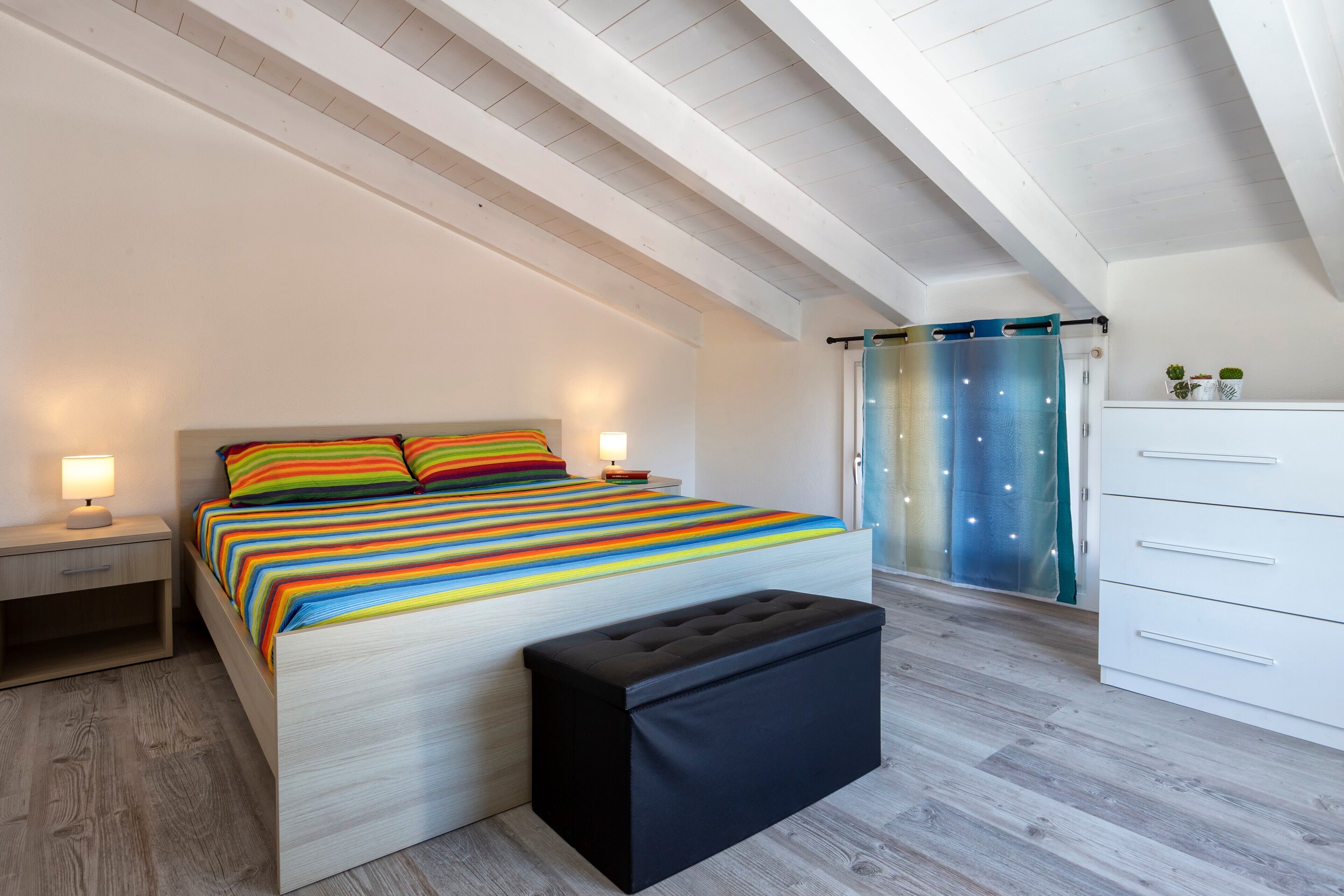 Ein Schlafzimmer mit einem Bett, Nachttischen, einer Bank, einem Schminktisch und einem Fenster mit Vorhängen.