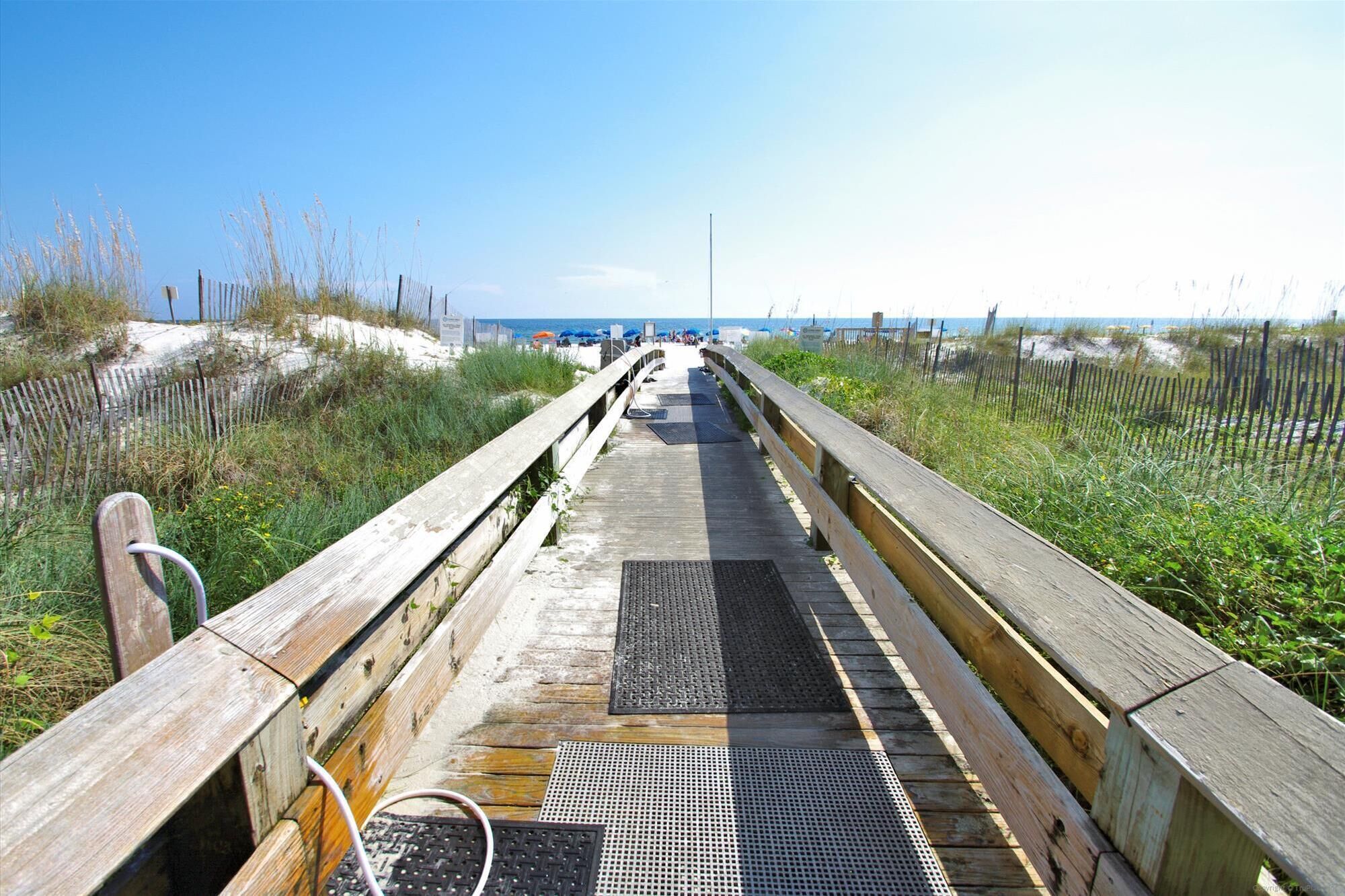 A wooden boardwalk leading to a beach with sand dunes and a clear sky.
