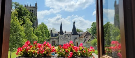 Vista através de uma janela de um edifício histórico com duas torres, cercado por vegetação exuberante e flores vibrantes.