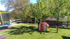 Ein grüner Außenbereich mit einem kleinen Spielhäuschen in Rot und Grün, einem Holzzaun und einem Steinhaus im Hintergrund.