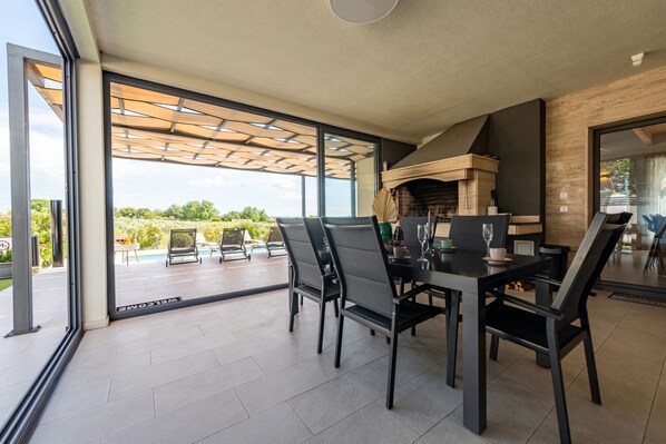 A modern outdoor dining area with a fireplace, large windows, and a view of the outdoors.