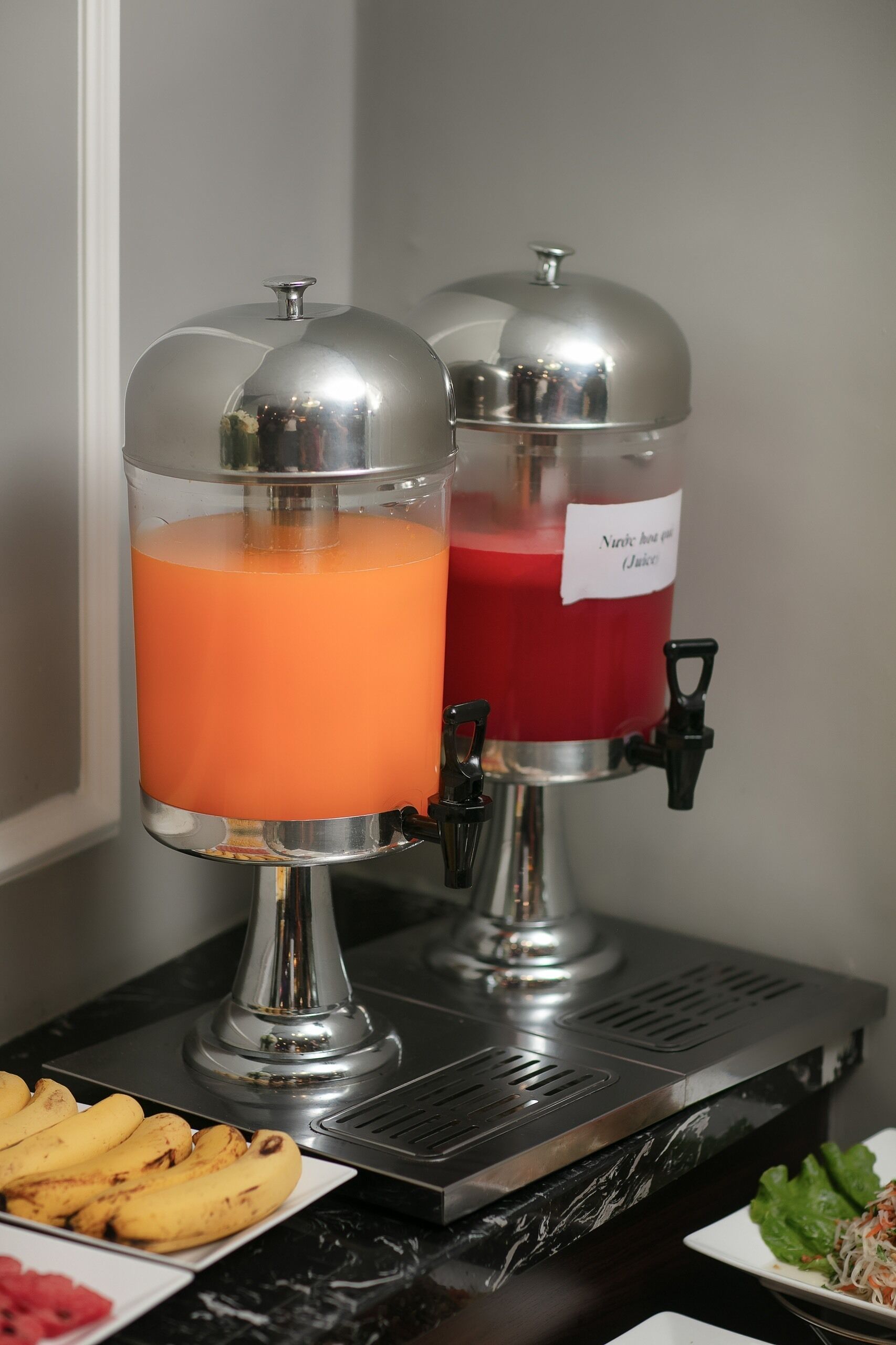 Two beverage dispensers on a counter with a plate of bananas and a salad.
