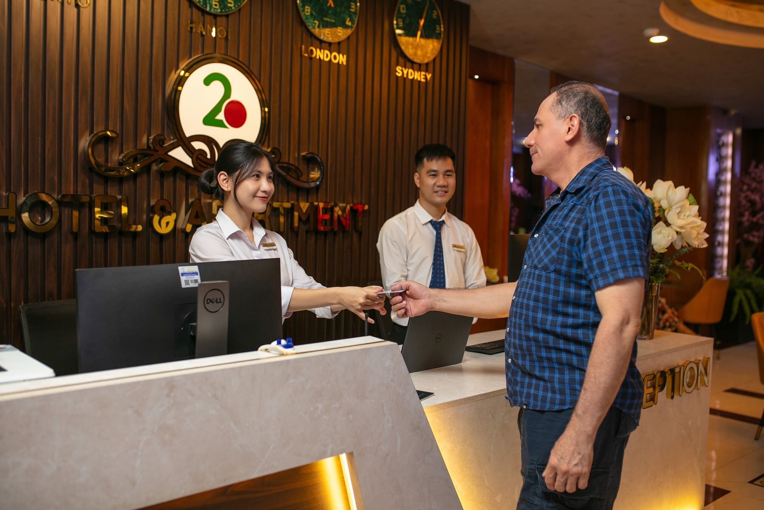Een hotel receptiebalie met een digitaal scherm, een balie en een muur waarop de naam van het hotel en stadsnamen staan.