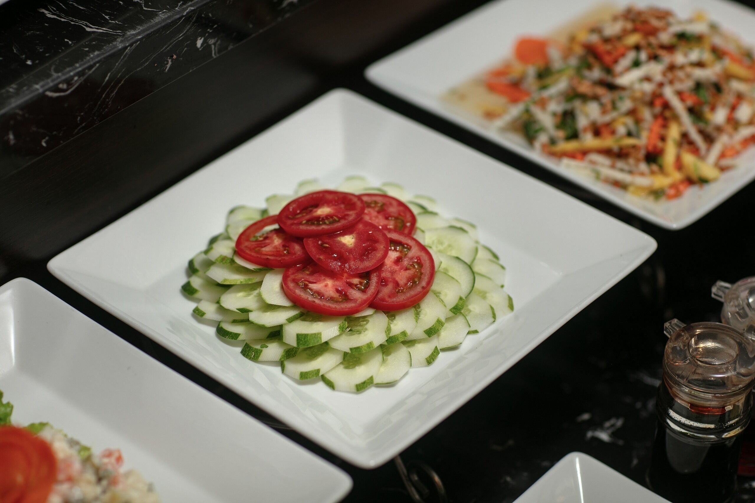 Ein Tisch mit verschiedenen Speisen, darunter ein Gurken-Tomaten-Salat, ein gemischter Gemüsesalat und ein Gericht mit geriebenem Käse.