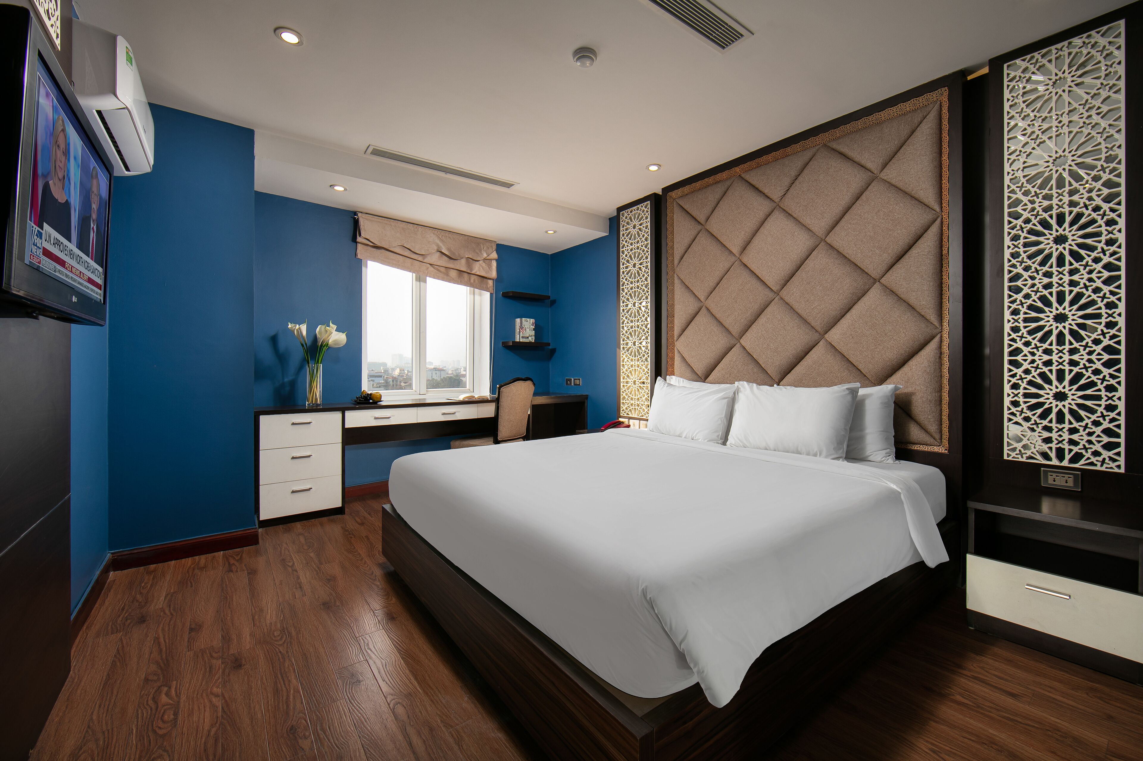 A modern hotel room with a large bed, a desk with a chair, a window with a view, and a decorative wall panel.