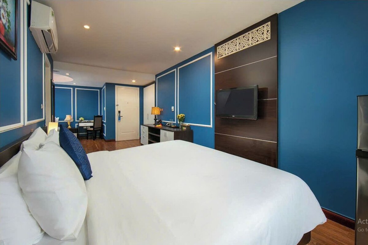 A hotel room with a large bed, a desk with a chair, a TV mounted on the wall, and a small kitchenette area.