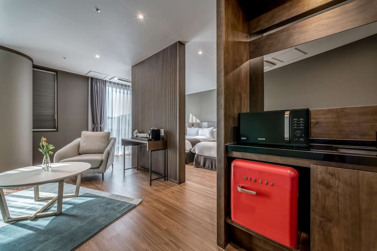 A modern hotel room with a wooden floor, a red minibar, a white sofa, a dining table with a vase of flowers, and a large window with curtains.