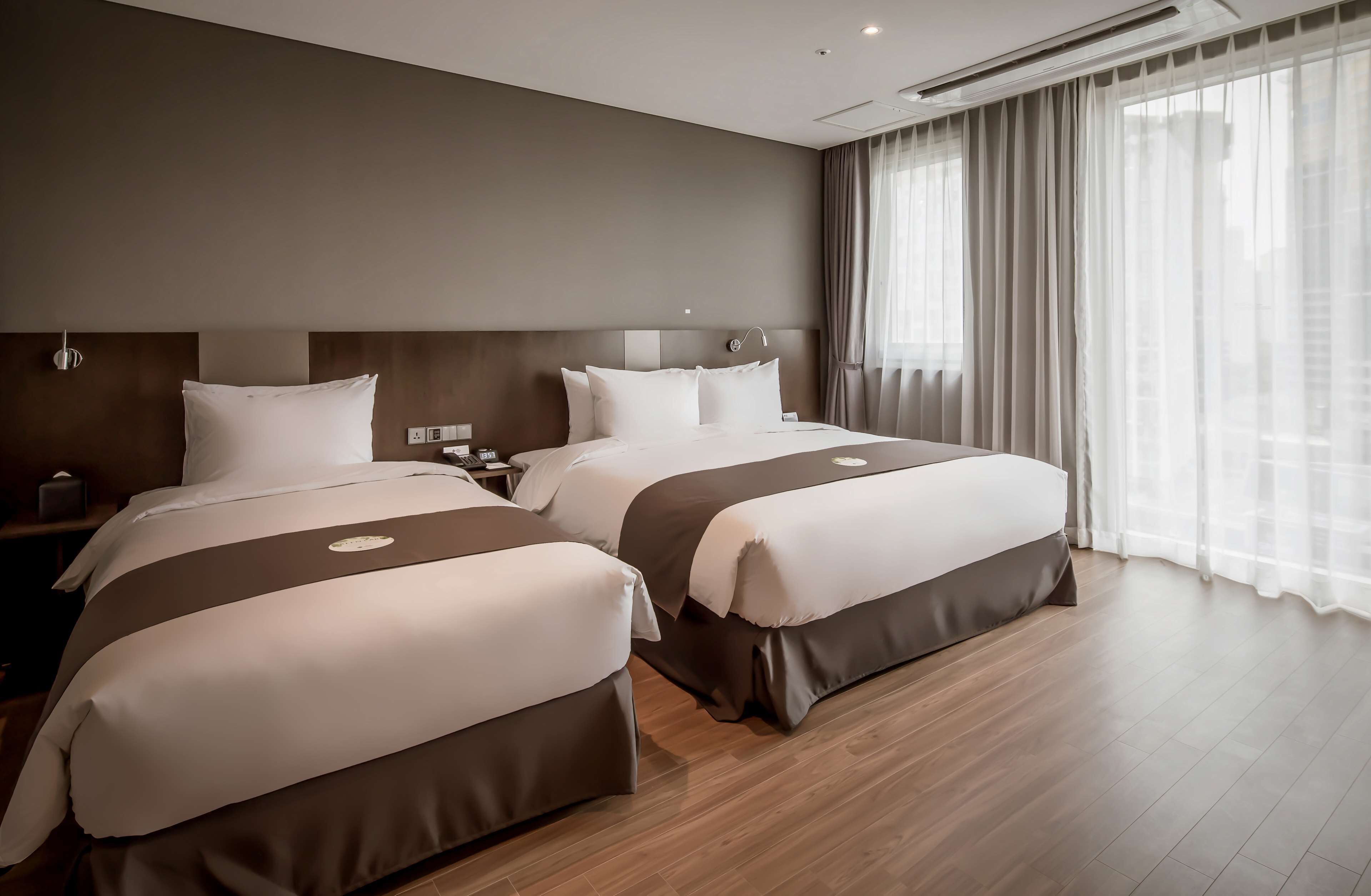 A hotel room with two beds, a wooden floor, a large window with curtains, and a modern headboard.