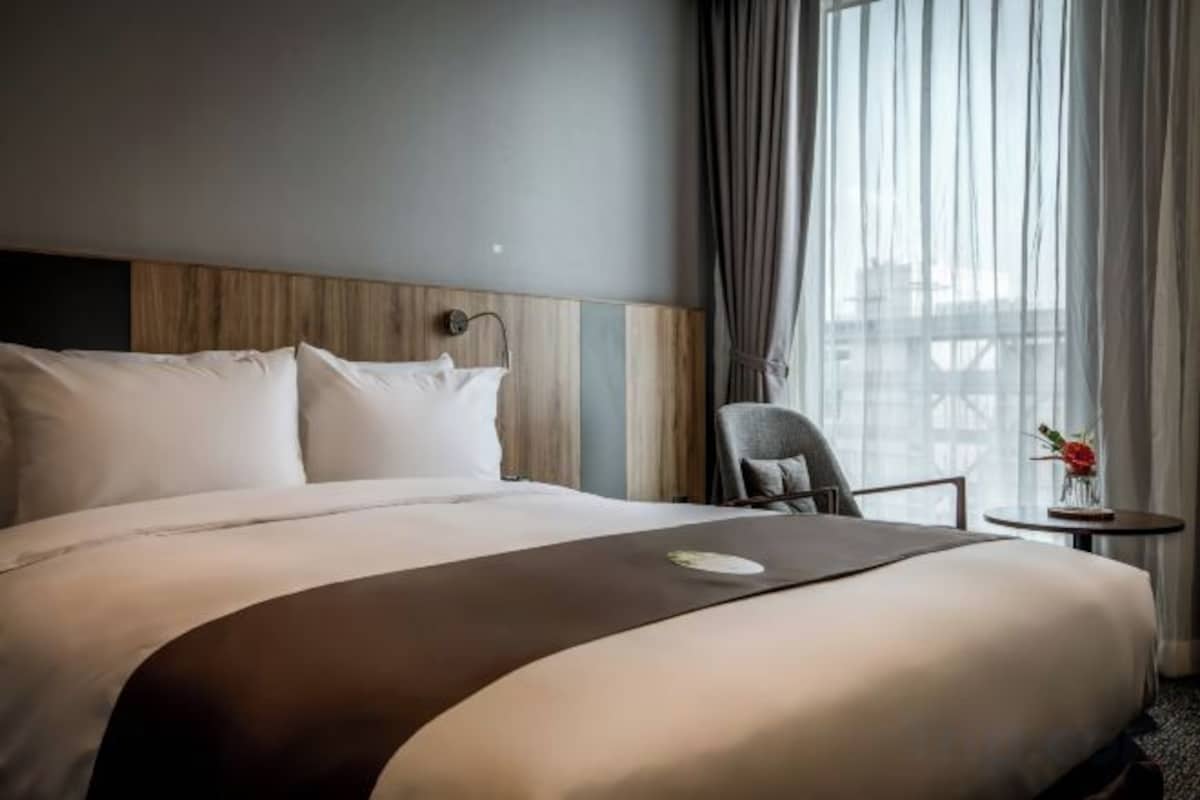 A hotel room with a large bed, a chair, a small table with a vase, and a view of a building through the window.