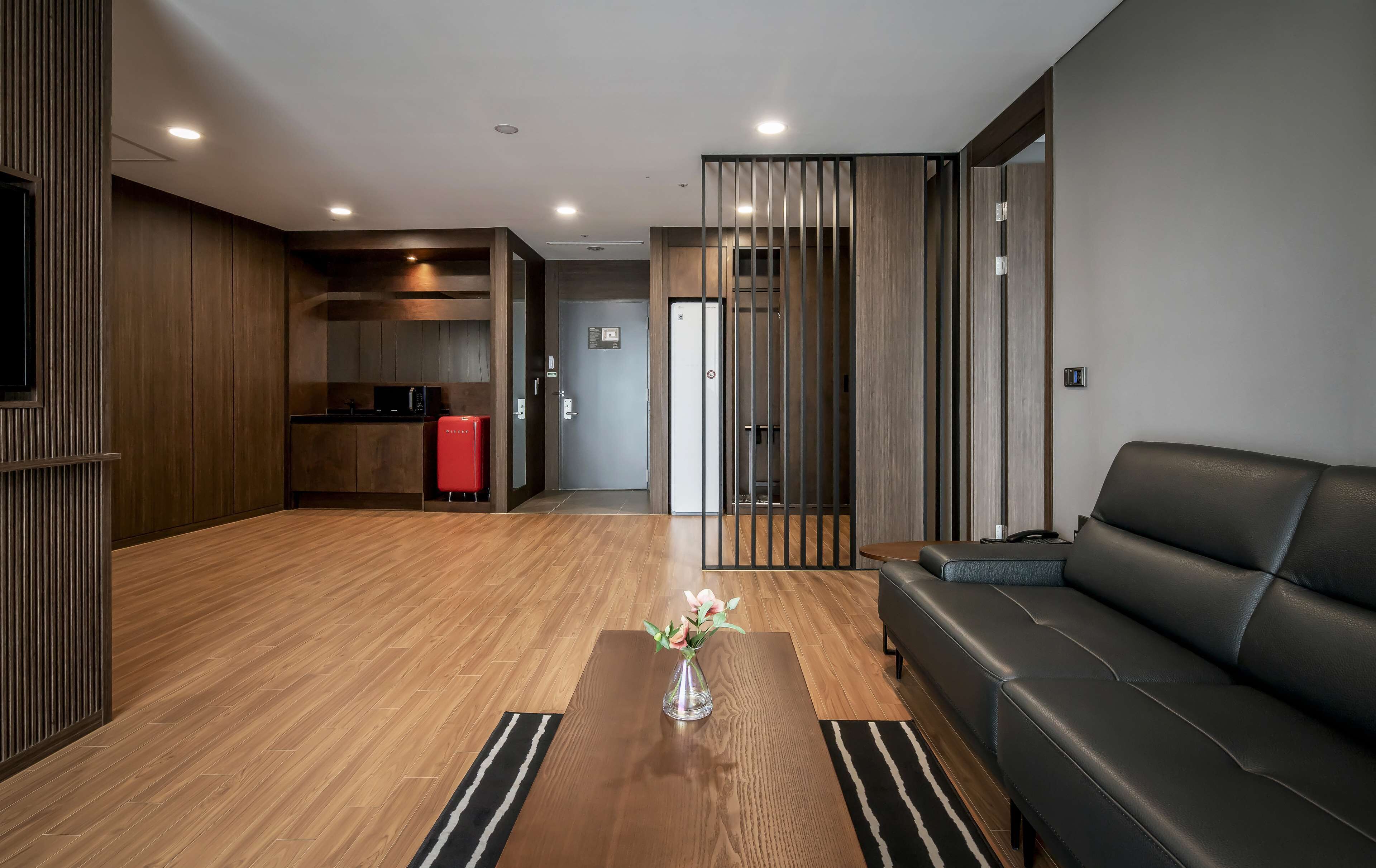 Una sala de estar moderna con un sofá de cuero negro, una mesa de centro de madera con un florero y una cocina con armarios de madera y un refrigerador rojo.