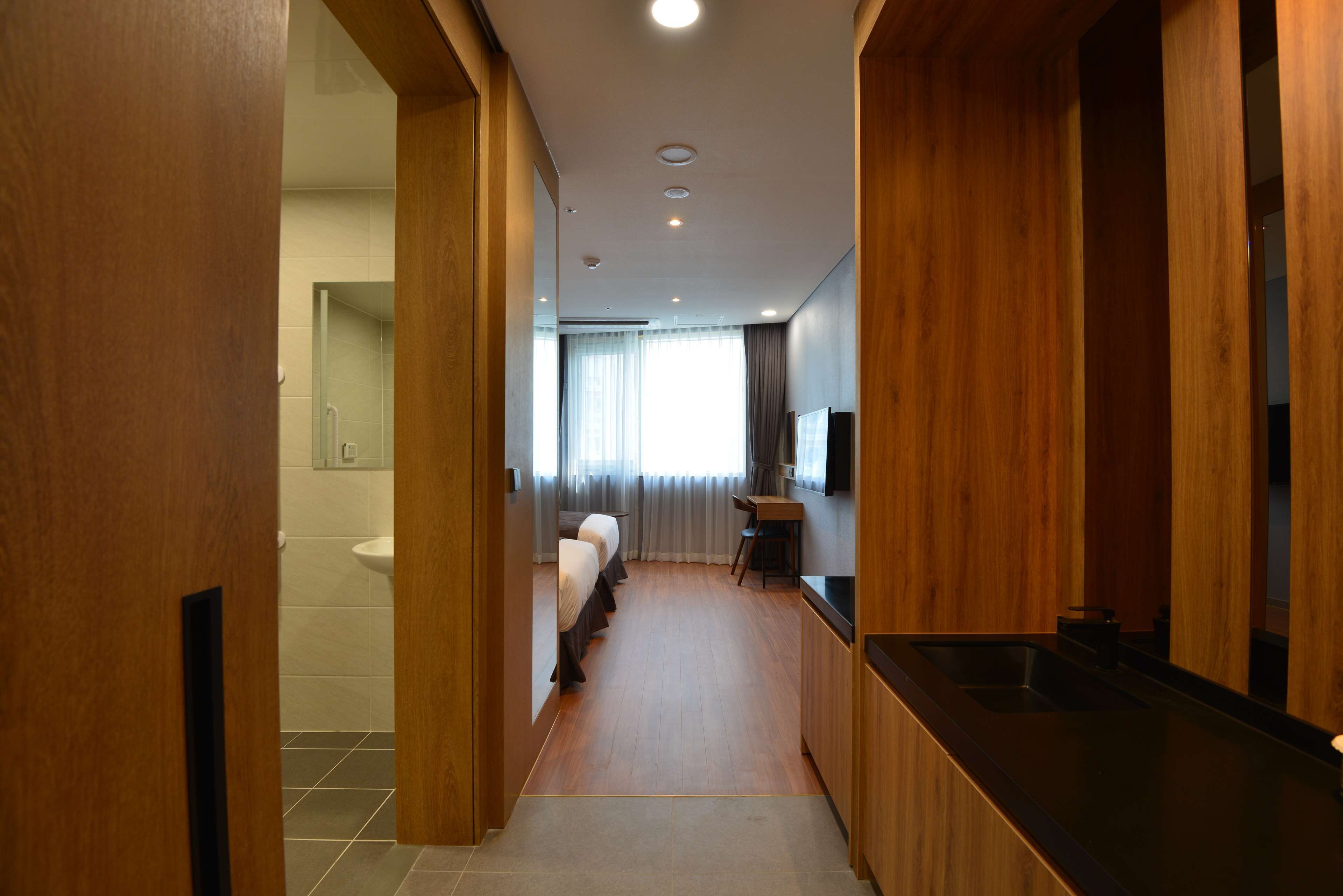 A hotel room with a wooden floor, a kitchenette, a bathroom with a toilet and sink, and a bed with a headboard.