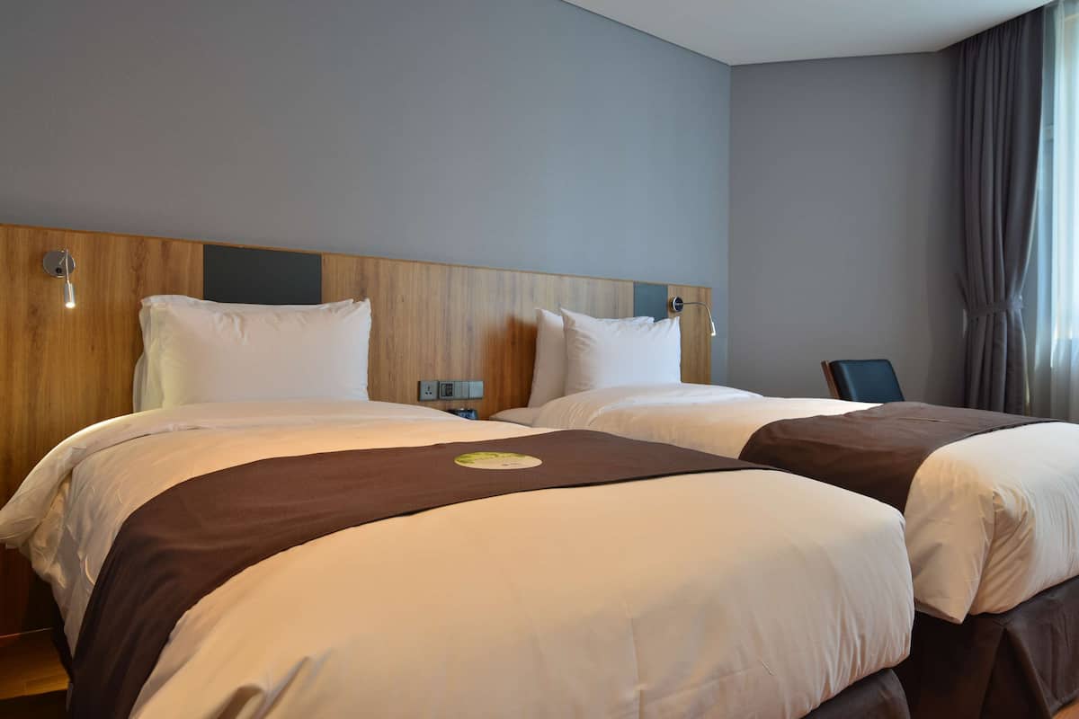 A hotel room with two beds, wooden headboards, a nightstand, and a view of the city.