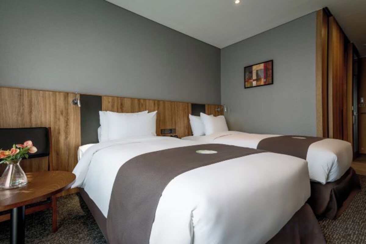 Quarto de hotel com duas camas, cabeceira de madeira, TV de tela plana e uma mesinha com um vaso.