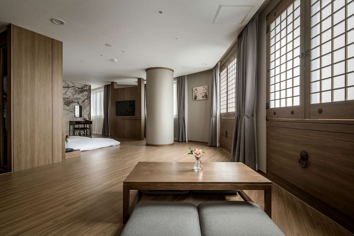 A modern living room with a wooden floor, a central column, a sofa, a coffee table with a vase, and large windows with curtains.