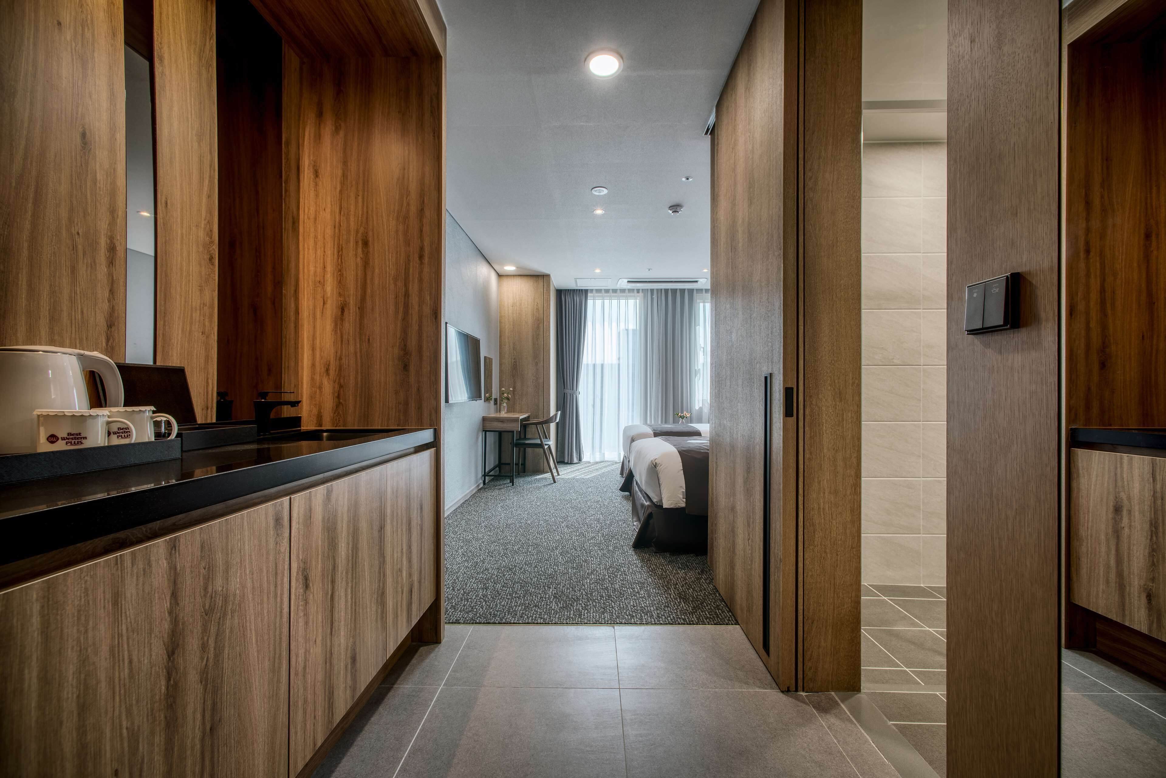 A hotel corridor with wooden paneling, a kitchenette, a bed, and a bathroom visible in the distance.