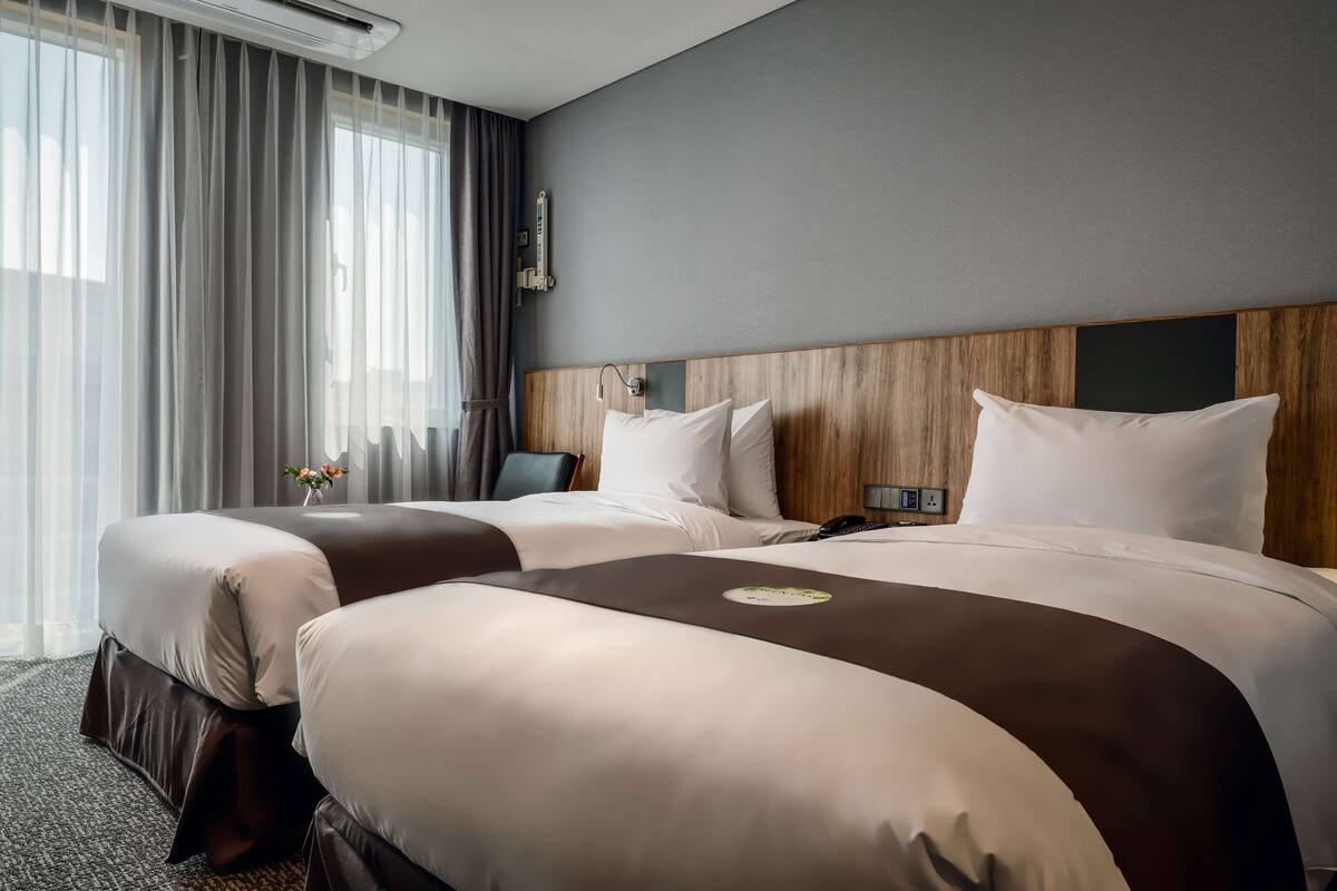 A hotel room with two beds, a wooden headboard, a bedside table, a lamp, and a window with curtains.