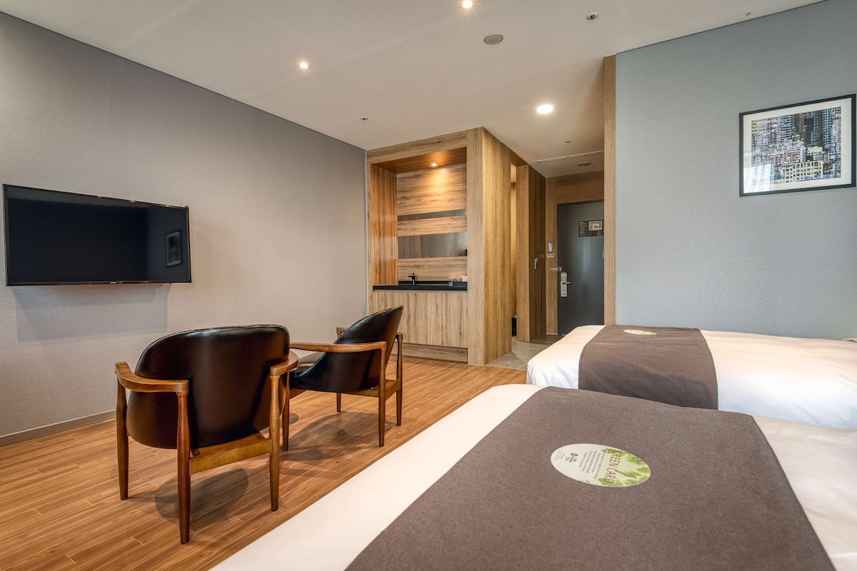 A hotel room with two beds, a wooden chair, a small table, a flat-screen TV, and a cityscape artwork on the wall.