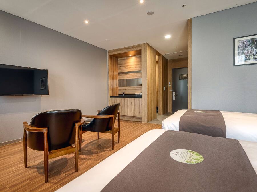 Une chambre d’hôtel avec deux lits, une chaise en bois, une petite table, un téléviseur à écran plat et une œuvre d’art représentant un paysage urbain accrochée au mur.