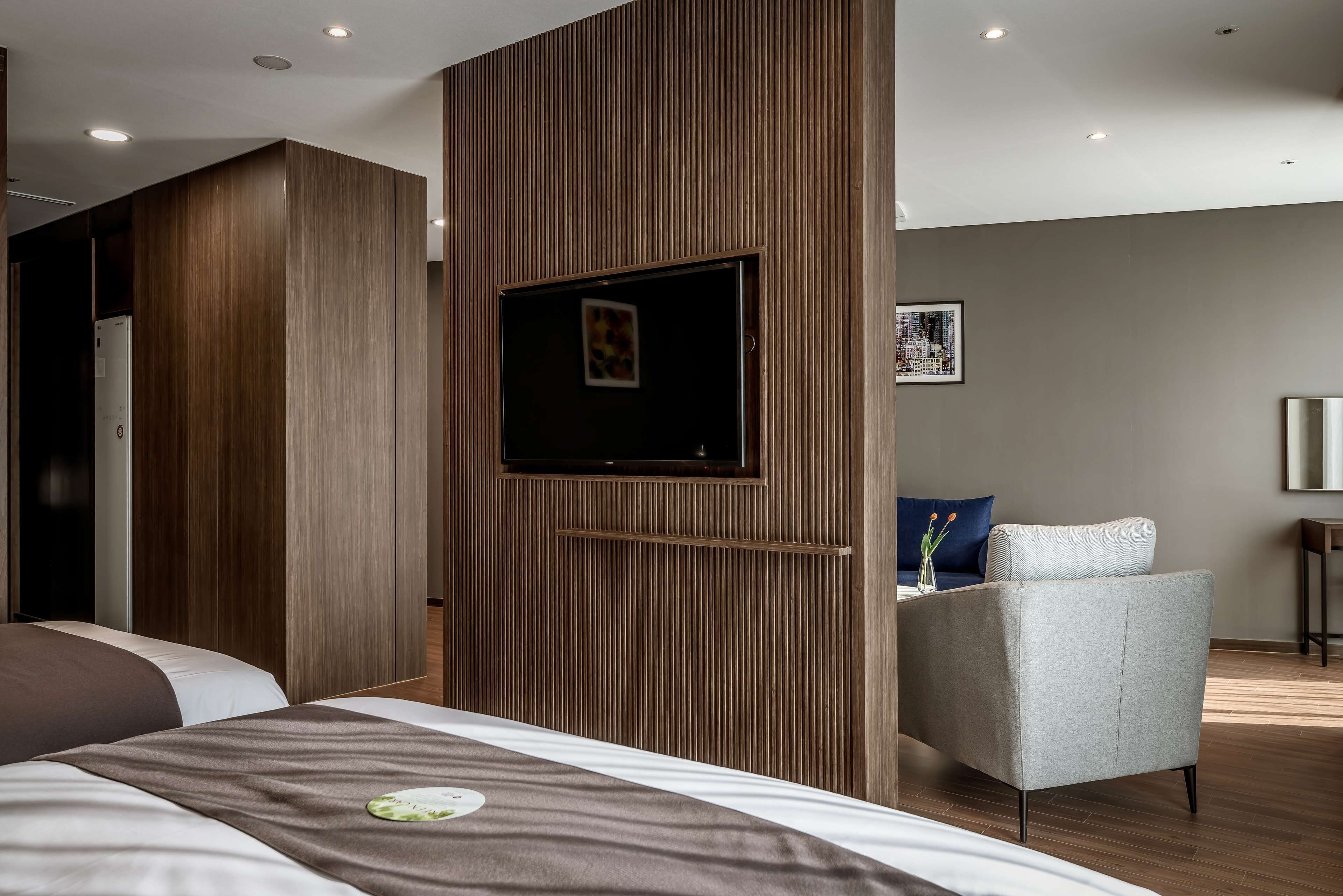 A modern hotel room with a large bed, a seating area with a chair, and a television mounted on a wooden panel.