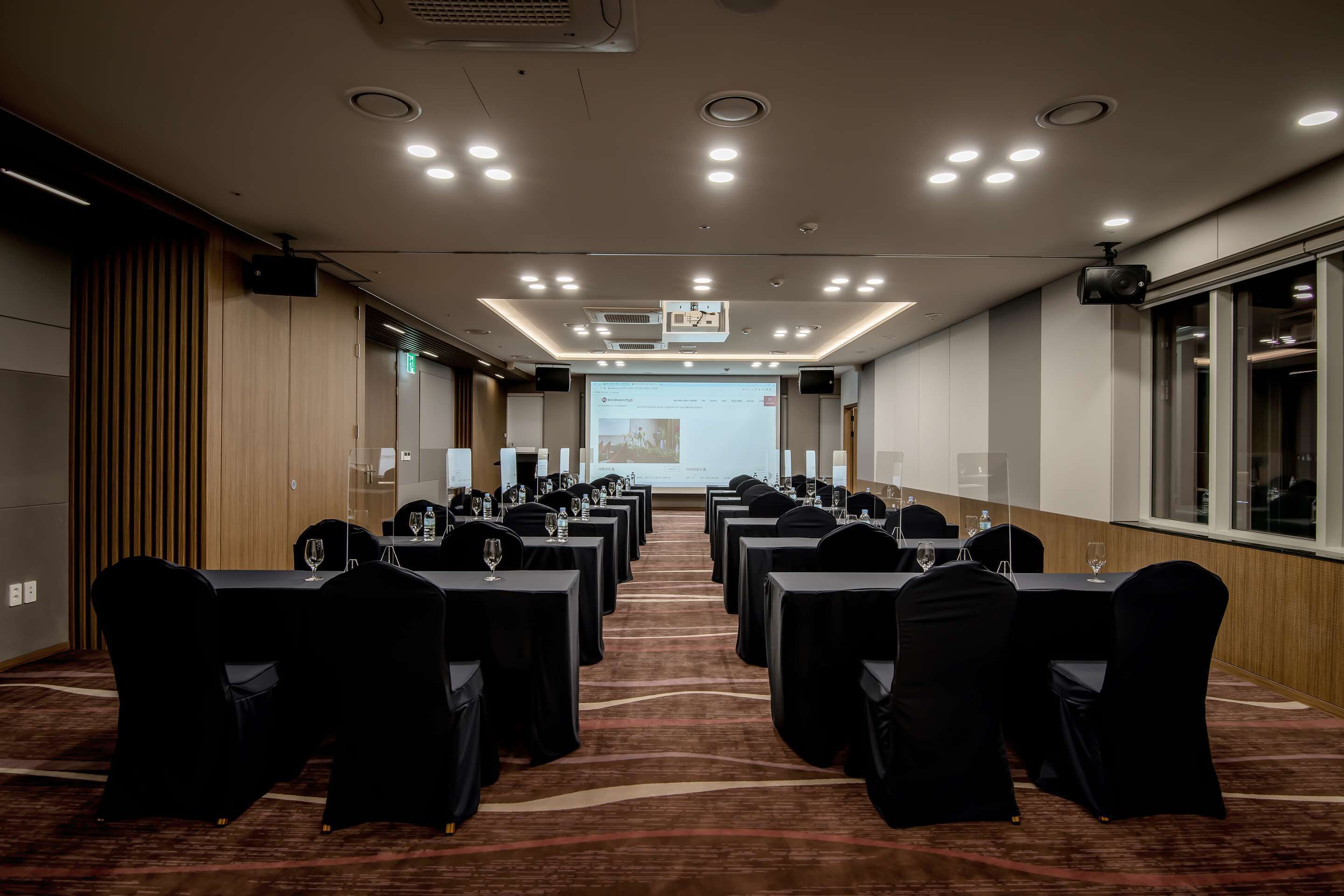 A conference room with rows of tables and chairs, a projector screen, and a carpeted floor.