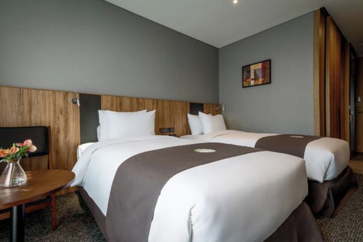 A hotel room with two beds, a wooden headboard, a small table with a vase of flowers, and a painting on the wall.