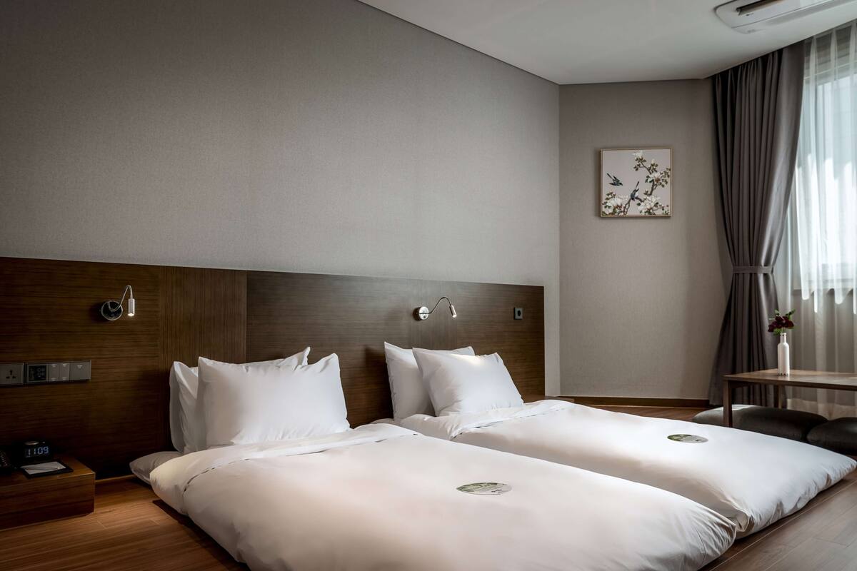 A hotel room with two beds, a wooden headboard, a bedside table with a lamp, a wall-mounted clock, and a framed picture.