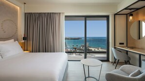 Habitación de hotel con cama, escritorio, silla, una mesita y un ventanal con vista al mar, además de una cortina.