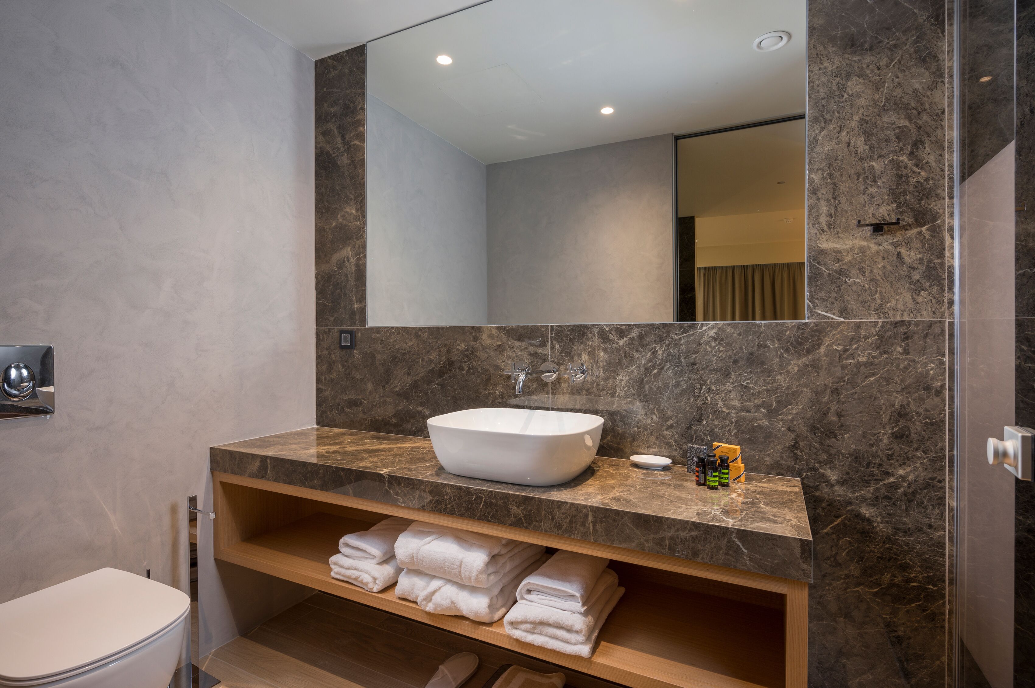 A modern bathroom with a large marble sink, a mirror, and a shelf with folded towels.