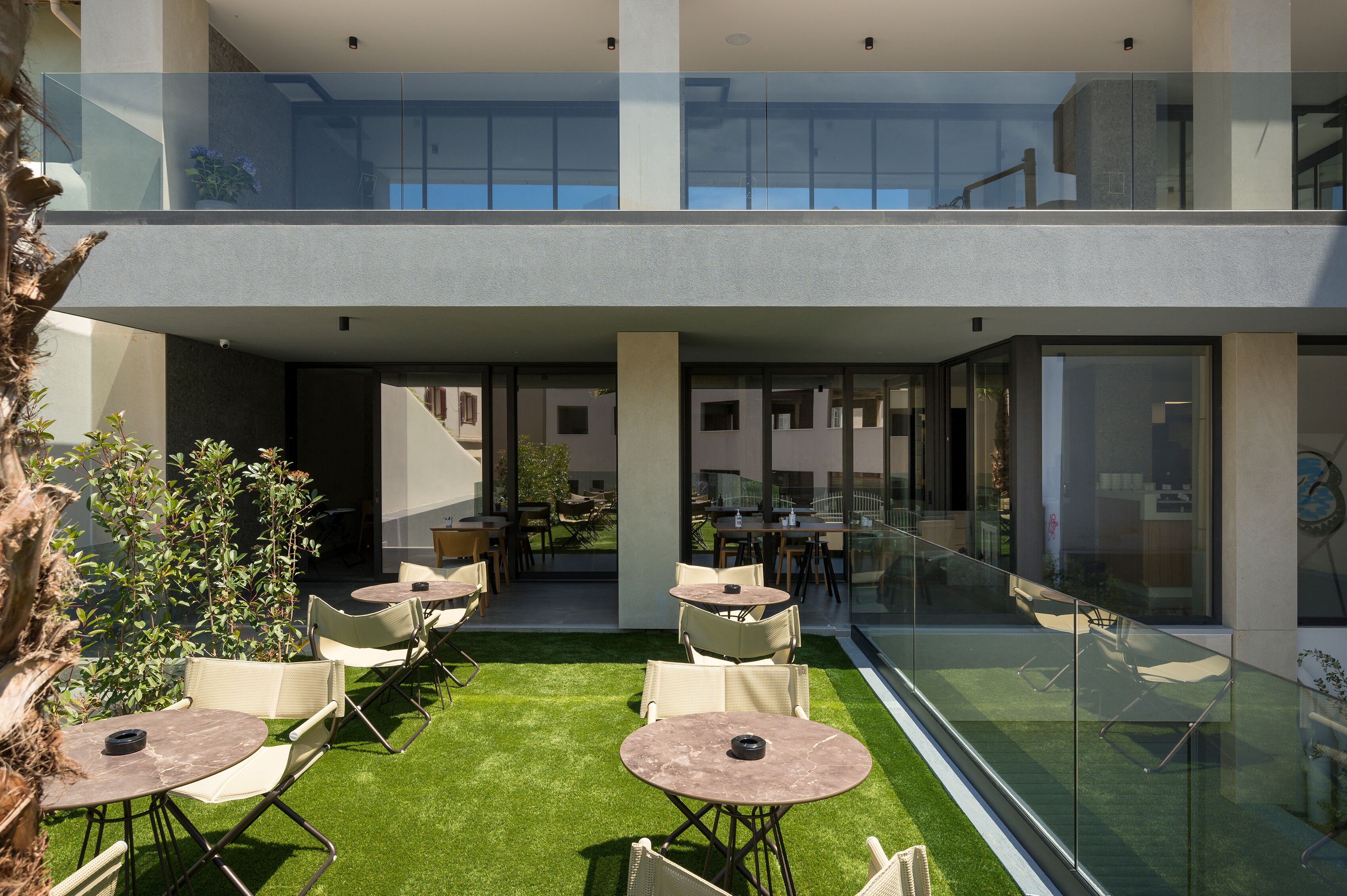 A modern outdoor seating area with tables and chairs, a glass railing, and a view of the interior.