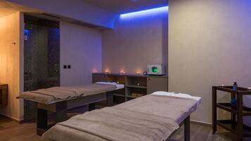 A spa room with two massage tables, a small shelf with candles, and a wall-mounted device.
