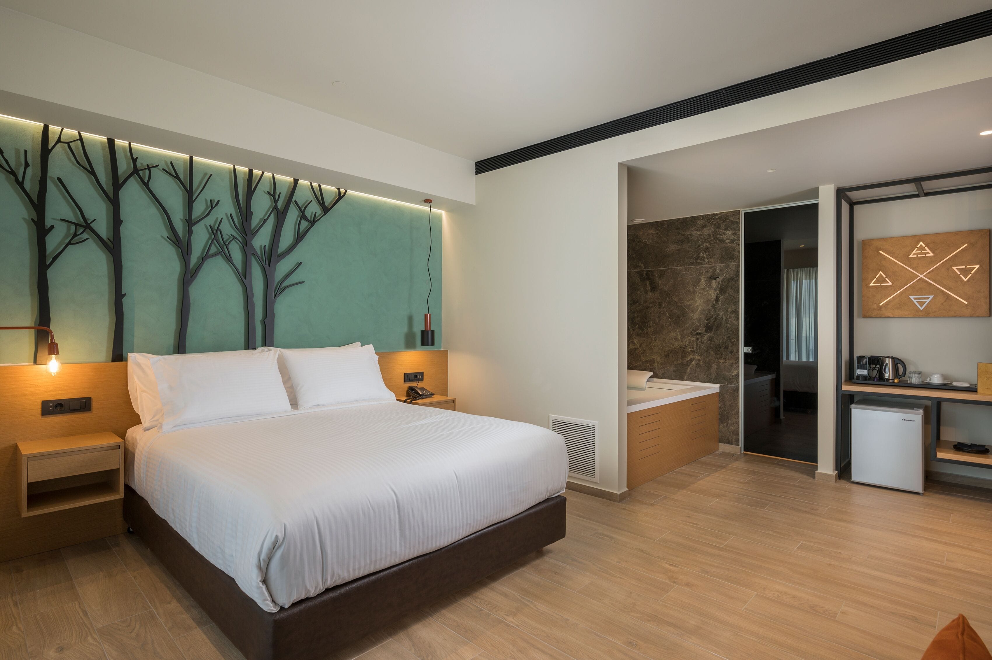 A modern hotel room with a large bed, a wall-mounted tree design, a small kitchen area, and a separate bathroom.