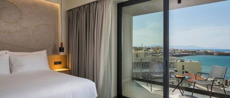 A modern hotel room with a large bed, a tablet on the bedside table, a view of the sea, and a balcony with a table and chairs.