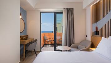 A hotel room with a bed, a desk, a chair, a small table, and a view of the ocean through a glass door.