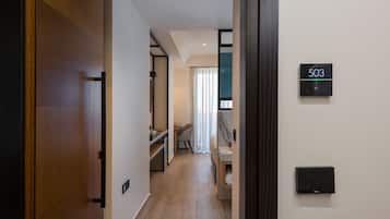 A hotel room with a wooden door, a digital lock, a small desk with a chair, a bed, and a bathroom with a towel rack.