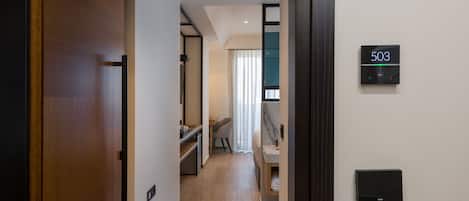 A hotel room with a wooden door, a digital lock, a small desk with a chair, a bed, and a bathroom with a towel rack.