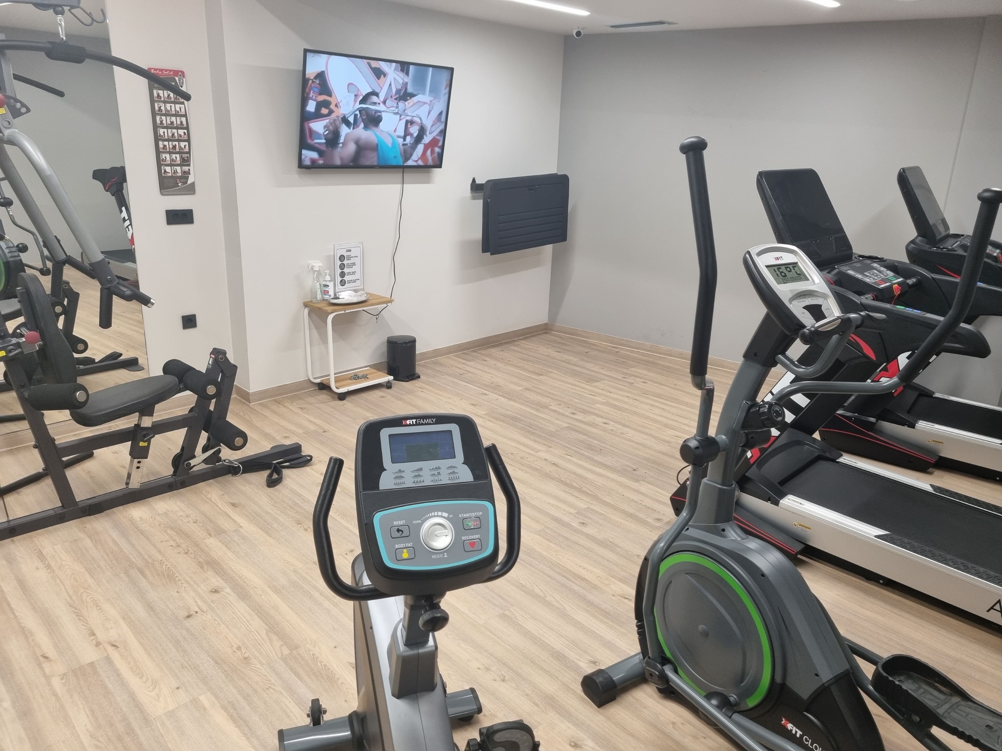 A gym with cardio equipment, a TV on the wall, and a small shelf with a water bottle.