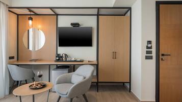 Une chambre d’hôtel moderne équipée d’un téléviseur à écran plat, d’un miroir rond, d’un bureau avec une machine à café, d’une chaise et d’une petite table sur laquelle se trouve un verre de vin.