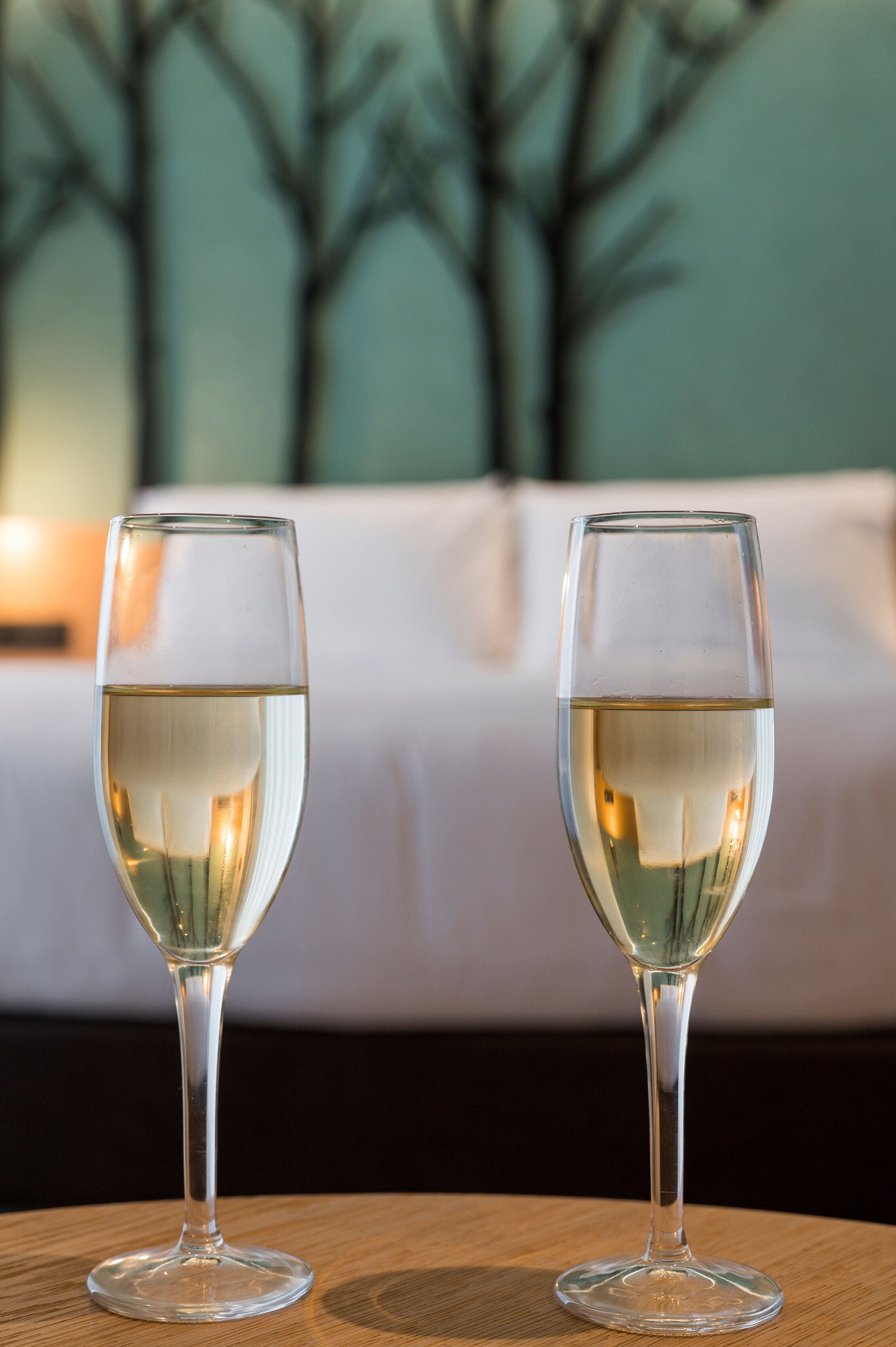 Two glasses of white wine on a round wooden table with a blurred background of a bed and a painting.