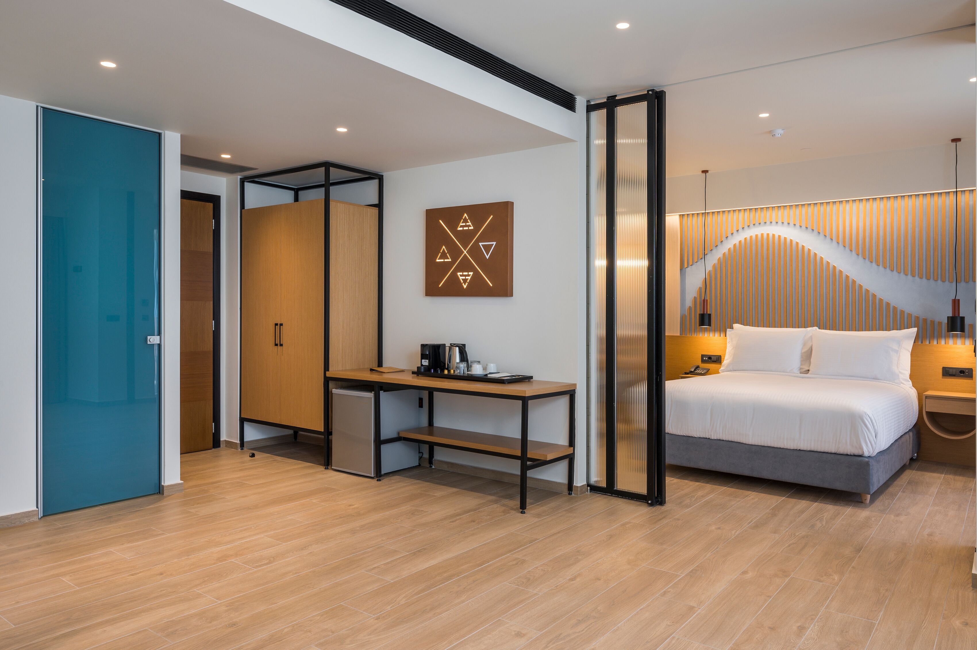 A modern hotel room with a wooden floor, a desk with a coffee maker, a bed with white linens, and a wall-mounted artwork.