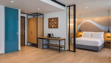 A modern hotel room with a wooden floor, a desk with a coffee maker, a bed with white linens, and a wall-mounted artwork.
