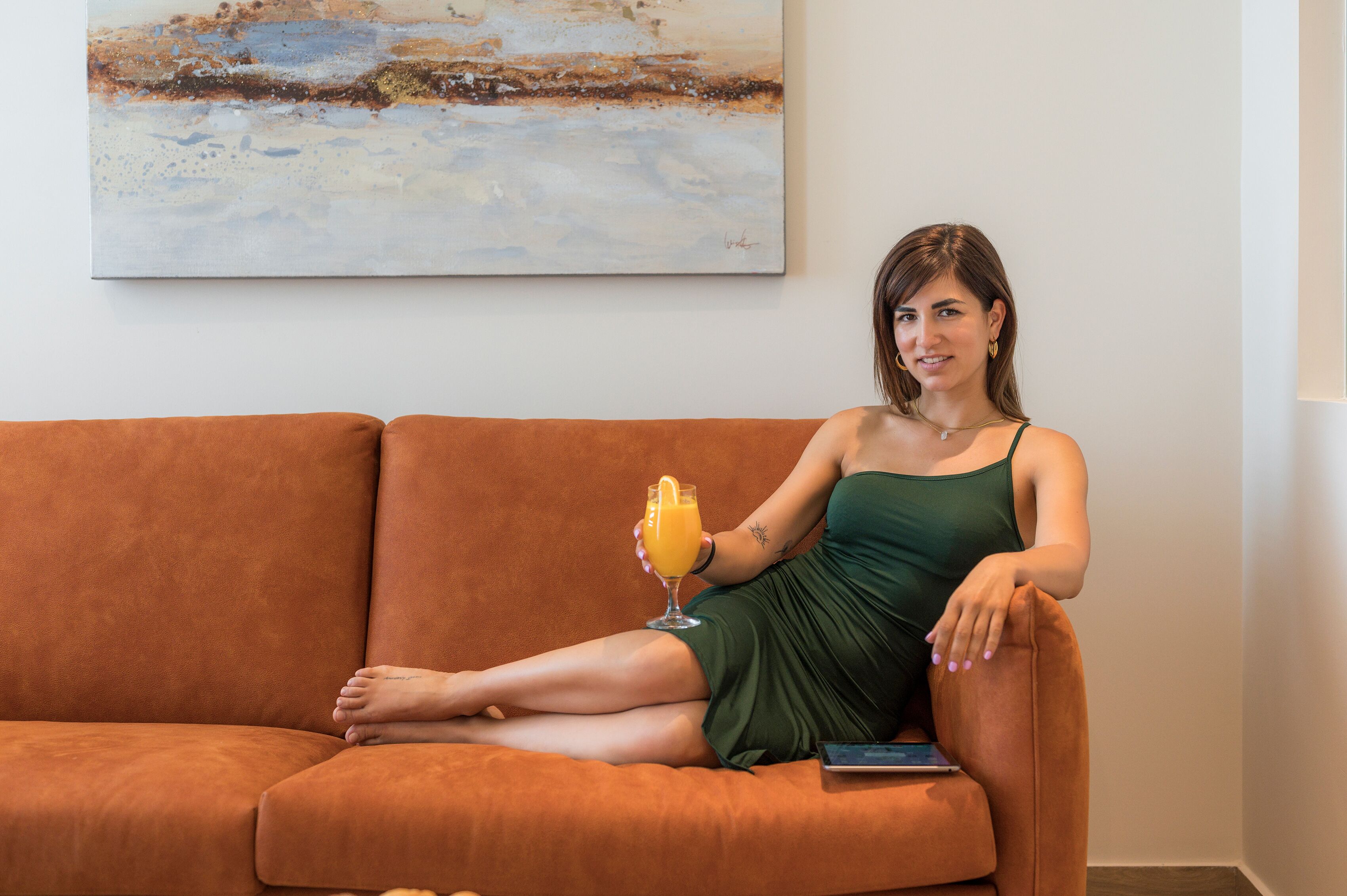 A person sitting on an orange sofa with a glass of orange juice, a painting on the wall, and a smartphone on the sofa.
