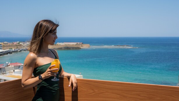 Una persona con un vestido verde sosteniendo un vaso de jugo de naranja, contemplando una vista costera con barandal de madera.