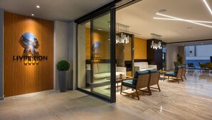 Un moderno vestíbulo de hotel con un mostrador de recepción de madera, un muro de cristal que da a una zona de estar con sofás y sillones, y una araña.