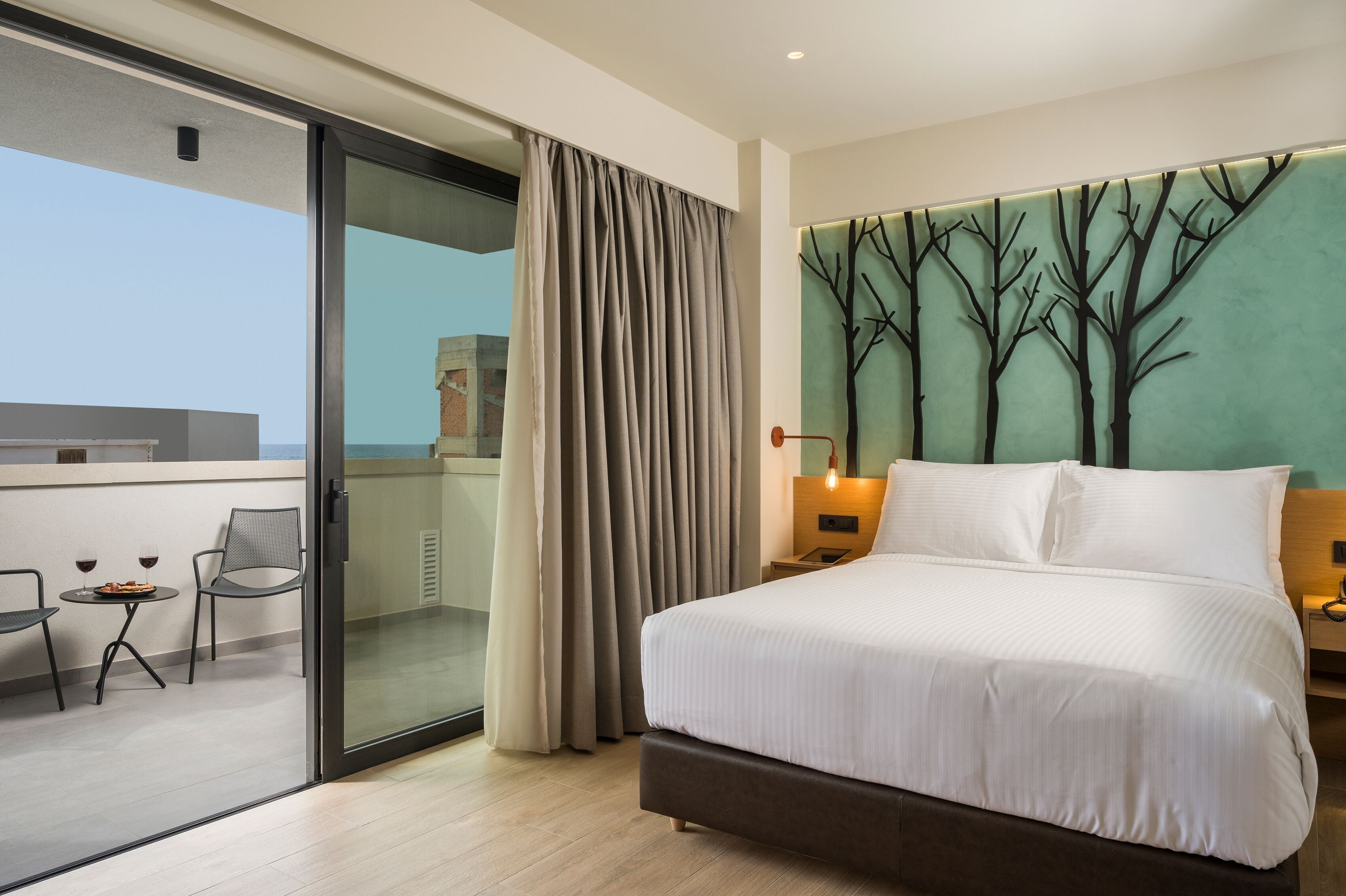 A hotel room with a large bed, a balcony with a table and chairs, and a wall with a tree mural.