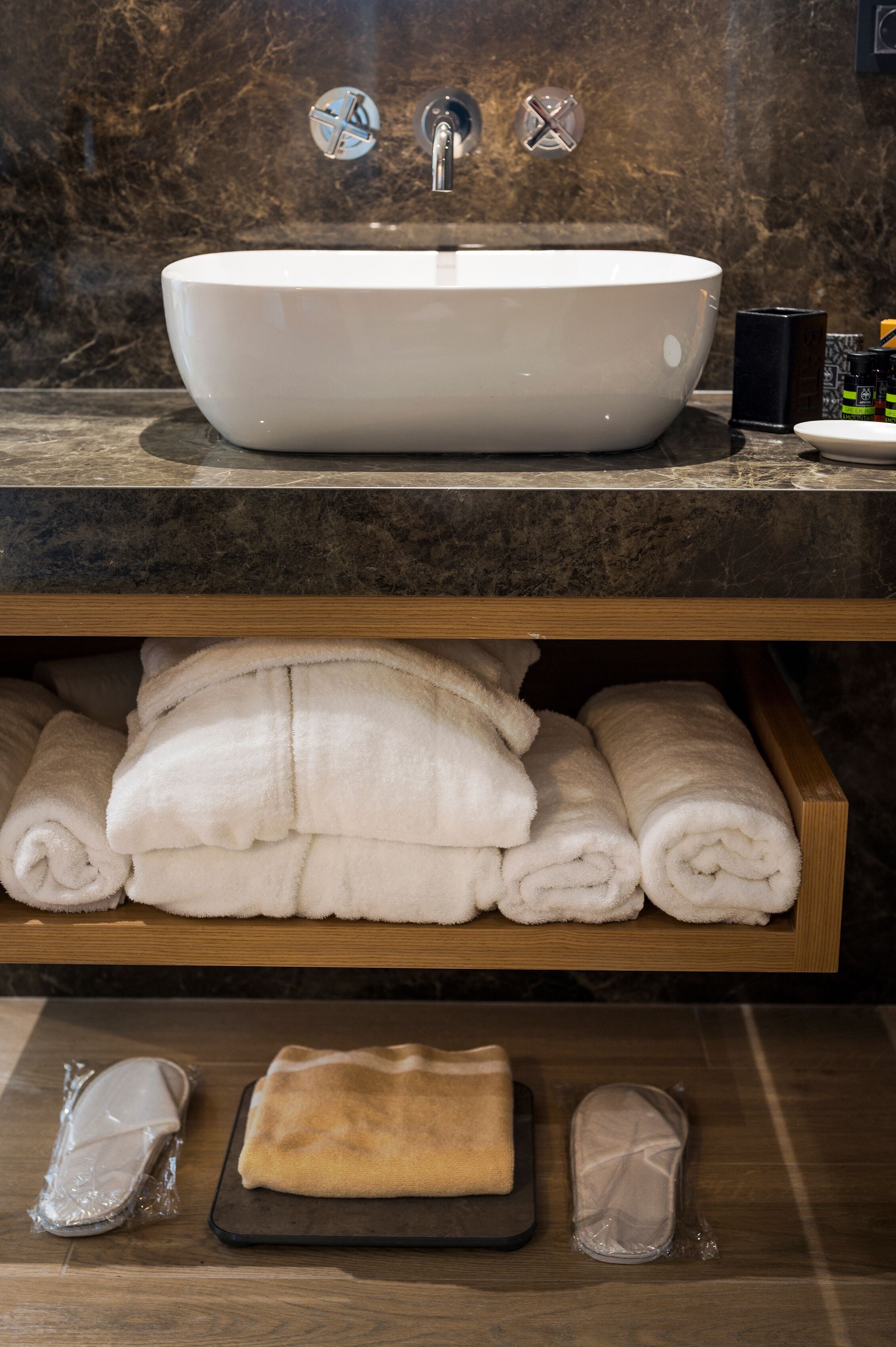 A bathroom with a large white sink, a wooden shelf with neatly folded towels, and a tray with a towel and two plastic bags.